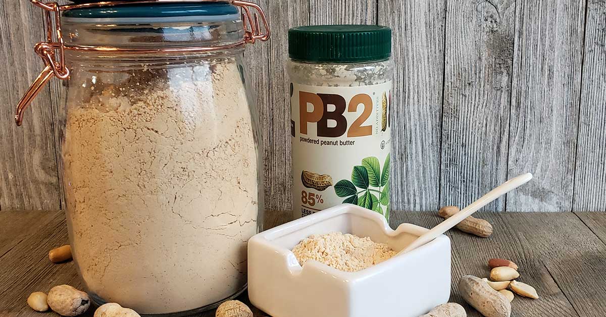 Glass jar with blue lid full of peanut powder with serving dish also full...all on a wooden background sprinkled with peanuts