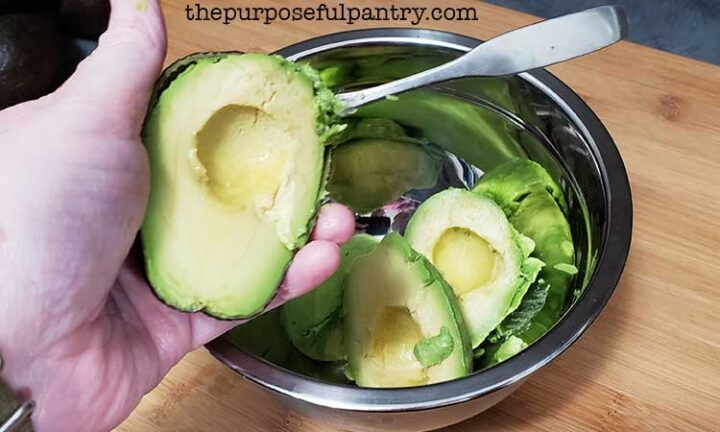 A halved avocado with spoon to remove the flesh over a bowl of avocados