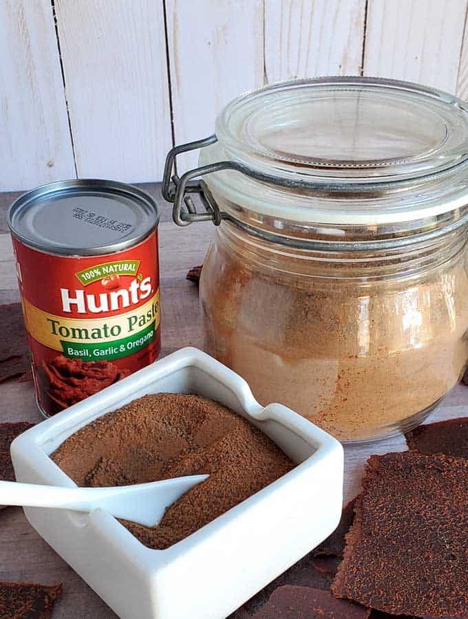 Tomato paste can, tomato powder in a glass jar and white serving dish with text overlay "How to Dehydrate Tomato Paste"