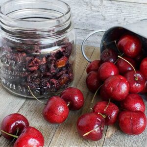 jar of dehydrated cherries alongside a silver serving dish of fresh cherries spilling onto a wooden surface