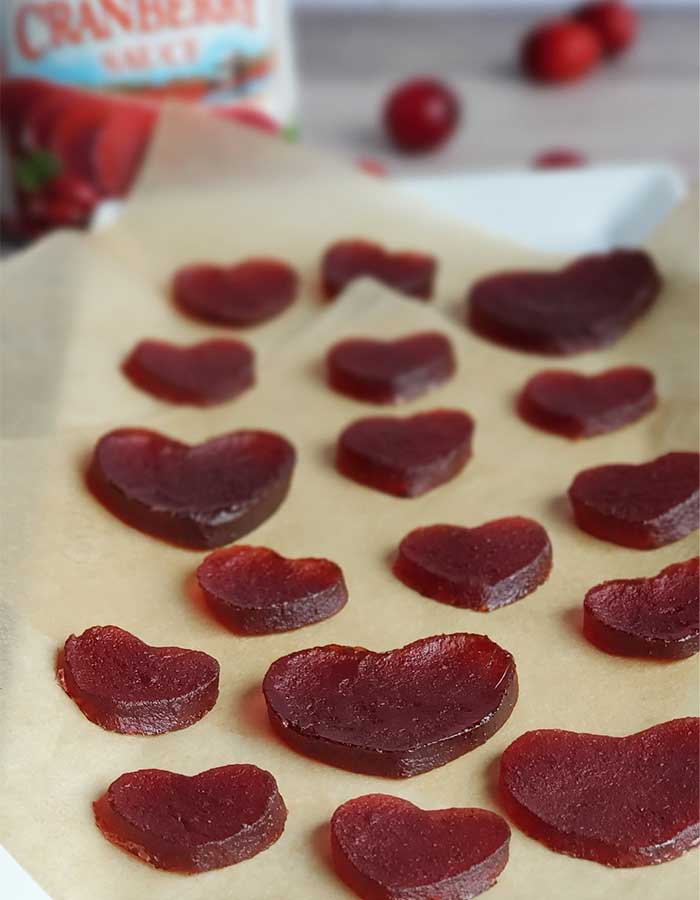 Dehydrated cranberry jelly heart gummies on parchment background with cranberries scattered and a can of cranberry jelly in the background.