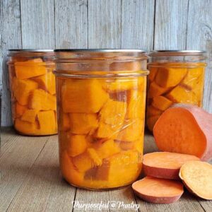 Mason jars of canned sweet potatoes with a fresh-cut up sweet potato all on a wooden backgroud
