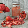 metal container with fresh strawberries, and two mason jars one with strawberry powder, one with dehydrated strawberries spilling out onto wooden surface