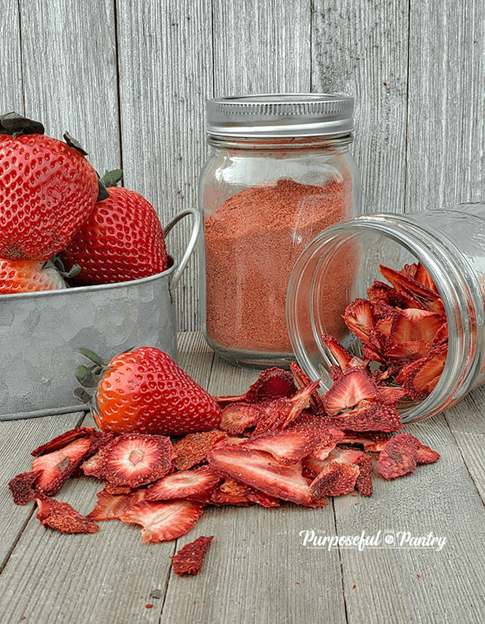 Fresh strawberries in a metal container with a jar of strawberry powder and a jar of dehydrated strawberries, all on wooden surface.