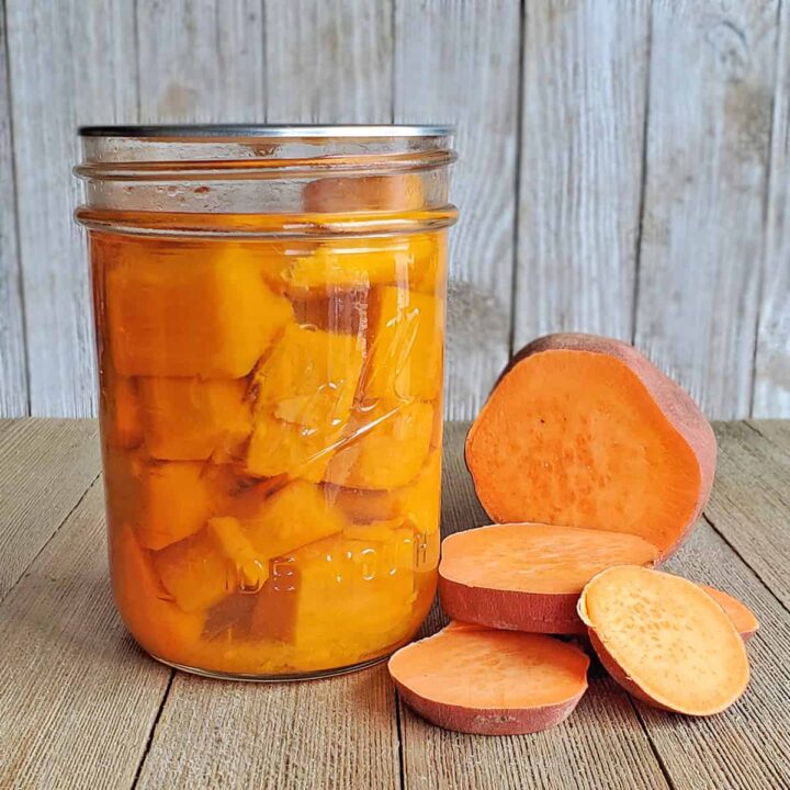 mason jars of canned sweet potatoes with a cut fresh sweet potato in the foreground all on a wooden surface