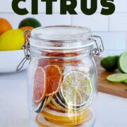 Glass jar full of dried citrus rounds on a wooden cutting board with cut lime, and whole lemon and orange