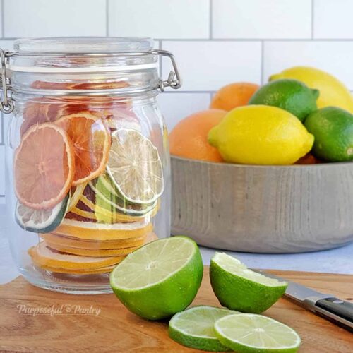 Jar of dehydrated citrus rounds in front of a bowl of fresh citrus, along with cut limes on a cutting board.