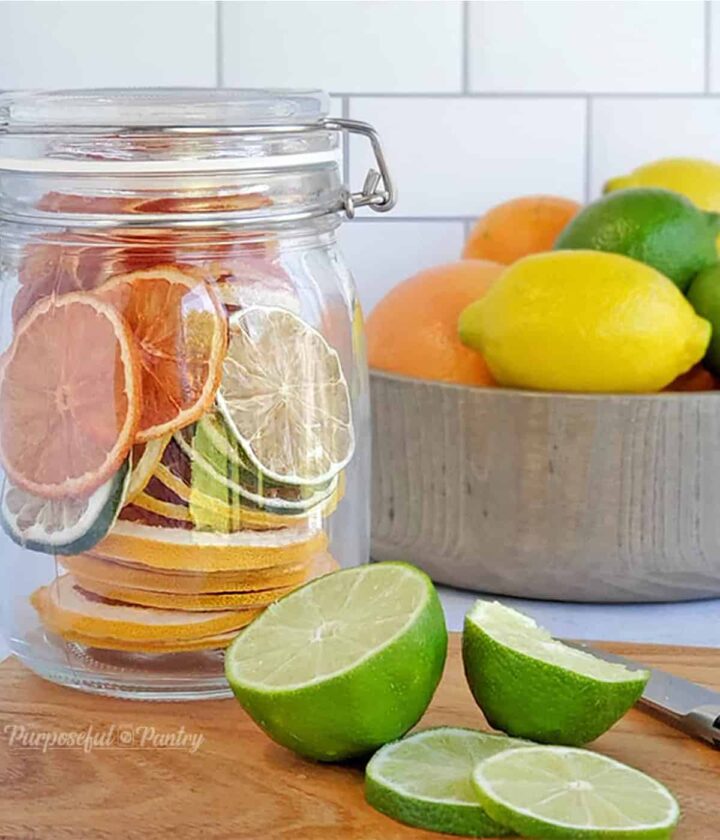 Jar of dehydrated citrus rounds in front of a bowl of fresh citrus, along with cut limes on a cutting board.