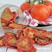 Dried tomato chips spilling onto a table from a jar, fresh tomatoes in the background