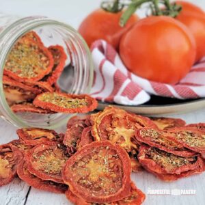 Dried tomato chips spilling onto a table from a jar, fresh tomatoes in the background