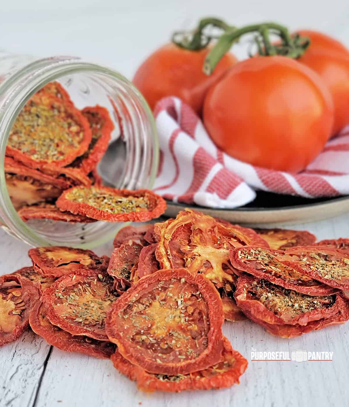 Dried tomato chips spilling onto a table from a jar, fresh tomatoes in the background.