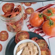 Pin Image fo garlic tomato spread on a blue plate with dried tomato slices and fresh tomatoes on a wooden surface