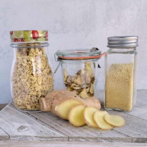 Jars of dried minced gigner, ginger slices, and ginger powder with fresh ginger laying on a white cutting board.