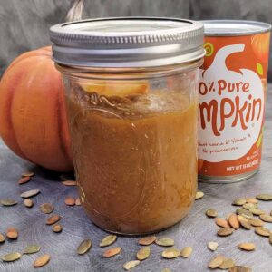 A canning jar of homemade pumpkin butter, with a can of pumpkin puree and pumpkin decor in the background, foreground sprinkled with pepitas