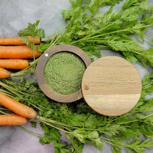 Carrot top powder in a wooden container surrounded by fresh carrots