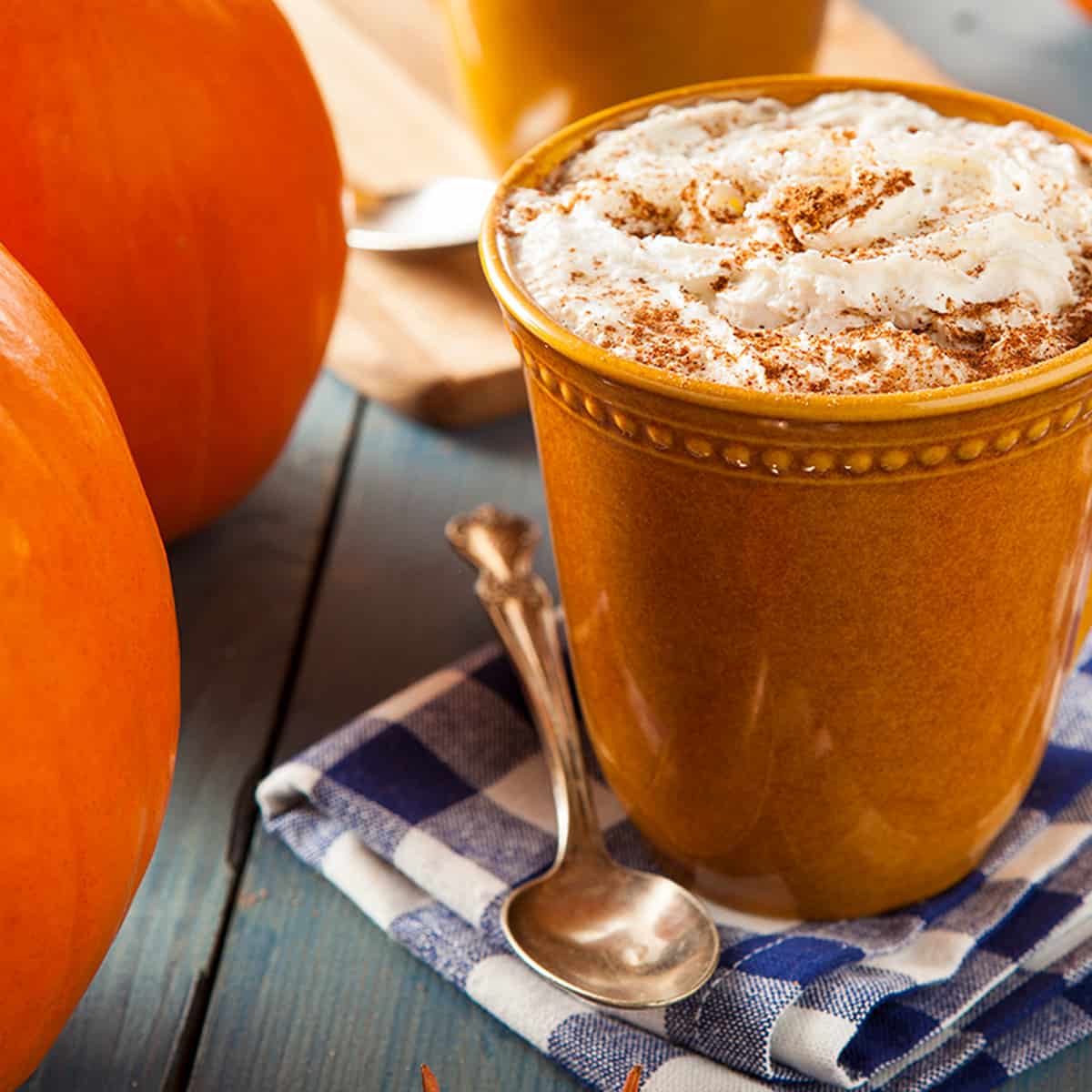 Orange coffee mug with pumpkins with pumpkin spice powder in foam on a blue checked napkin