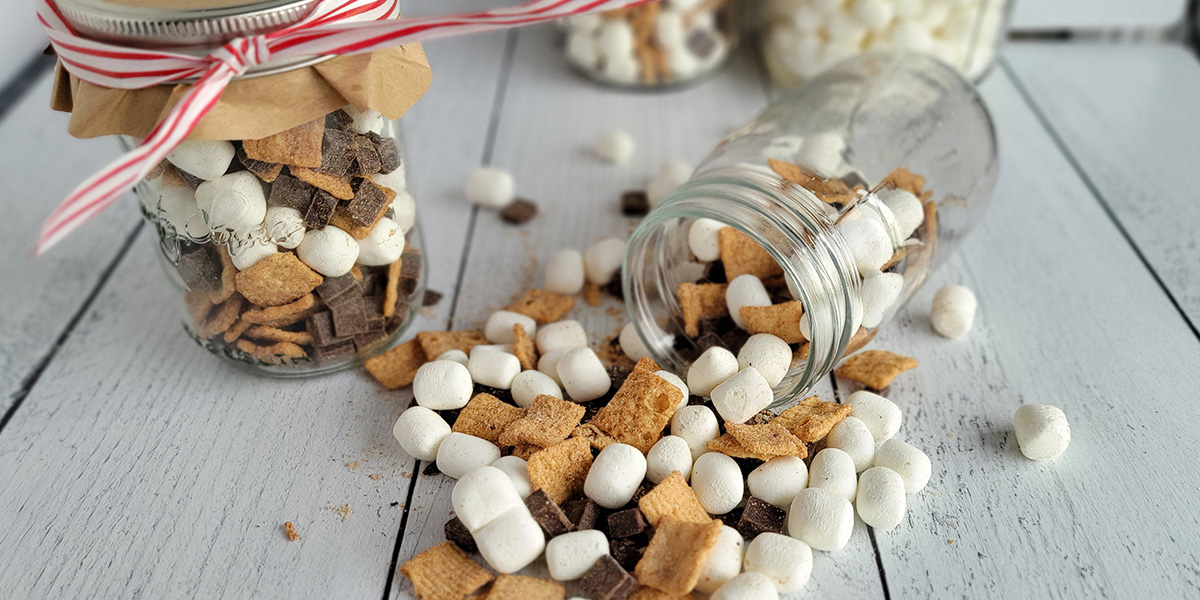 Crunchy s'more mix with dehydrated marshmallows, graham cereal and chocolate chunks in glass jars.