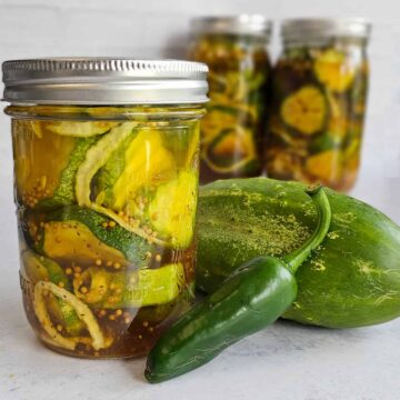 Canning jar of sweet and spicy pickles with a jalapeno and cucumber on a white background; pickle jars in backround.