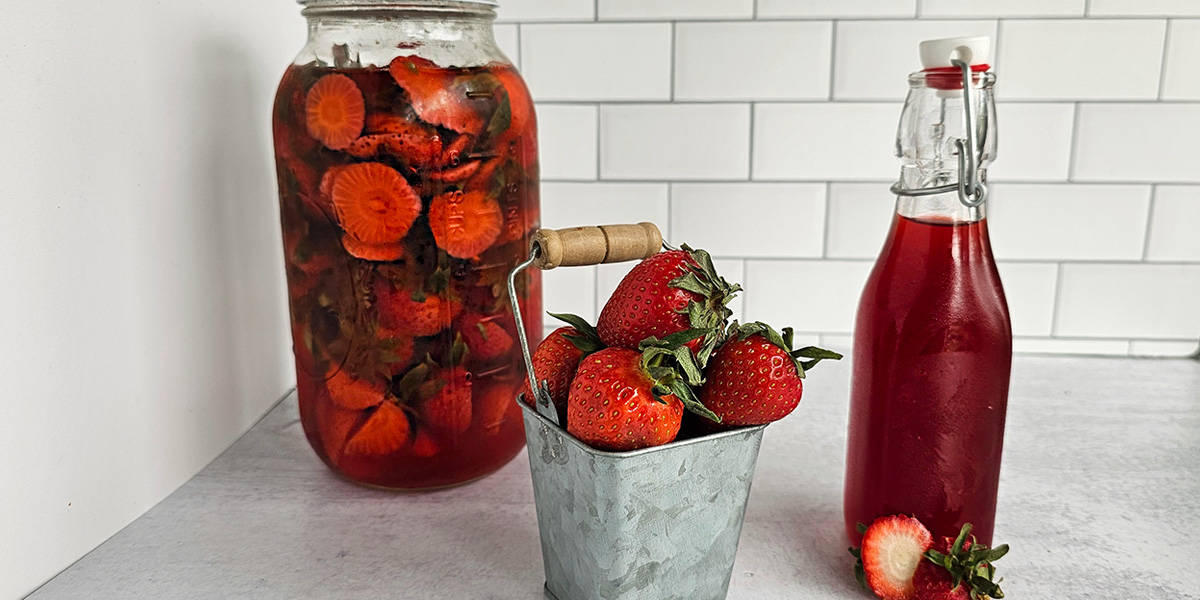 A large jar of strawberry-infused vinegar, a stack of strawberries in a metal tin, and a bottle of strawberry syrup on a white background.