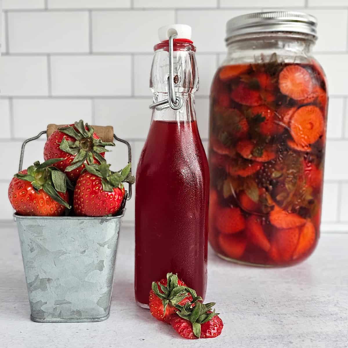 Bottle of strawberry syrup, A stack of strawberries in a metal container, a bottle of strawberry syrup, and a jar of strawberry-infused vinegar.