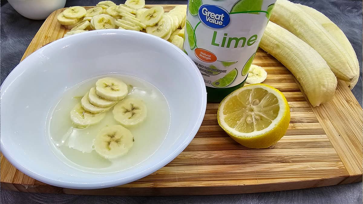 A bowl with bananas, lemon, and a bottle of lime juice on a wooden cutting board.