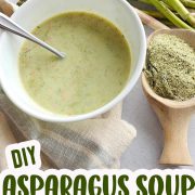 A bowl of homemade asparagus soup with fresh asparagus and a spoonful of dried herbs next to it.
