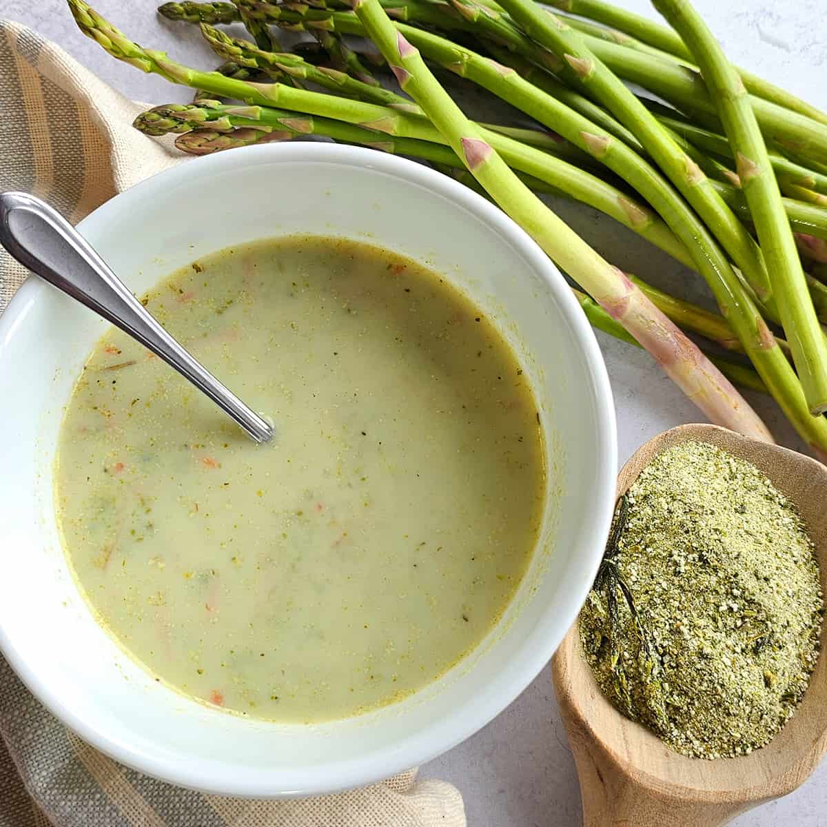 A bowl of creamy asparagus soup next to fresh asparagus stalks and a scoop of asparagus soup powder.