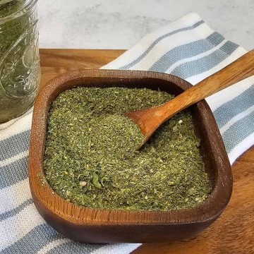 A wooden bowl filled with finely ground green herbs, with a small wooden spoon inside. A glass jar and a striped cloth are in the background.