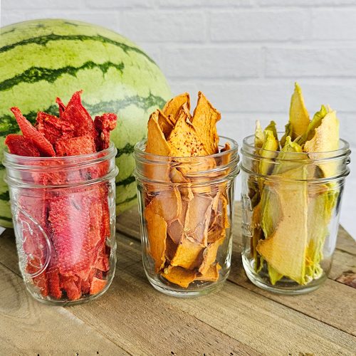 Three glass jars filled with dried watermelon, cantaloupe and honeydew melon are placed in front of a whole watermelon on a wooden surface with a white brick wall in the background.