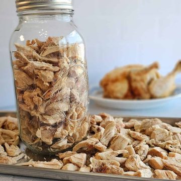 A jar filled with dried ginger pieces surrounded by more dried ginger on a tray, with a blurred plate of ginger in the background.